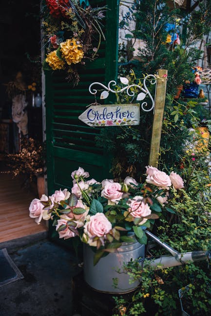 A floral display outside a florist shop features a lush arrangement of soft pink roses with velvety petals and green foliage, placed in a silver watering can. In the background, there are vibrant yell