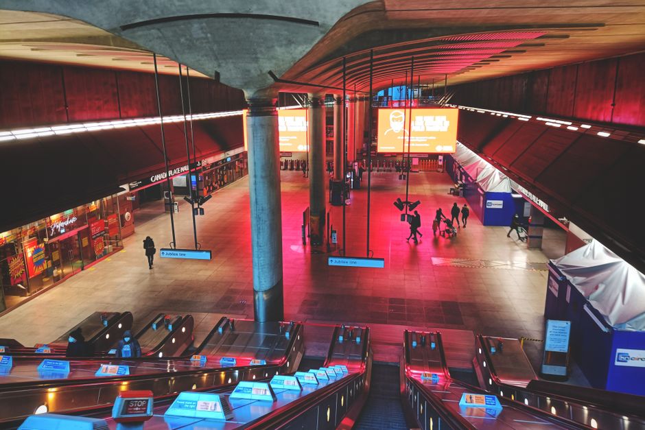 The image depicts an interior view of Canary Wharf station illuminated with warm, reddish lighting. The spacious platform features smooth, polished floor tiles and is lined with digital signage and in