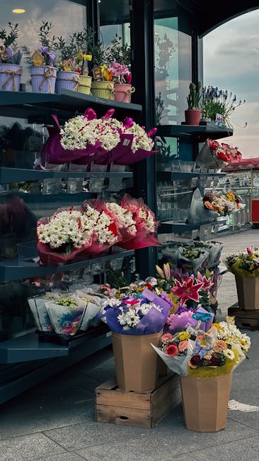 An outdoor display at a flower shop showcasing a variety of fresh floral arrangements. There are multiple bouquets wrapped in colorful paper and cellophane, featuring vibrant pinks, whites, purples, a