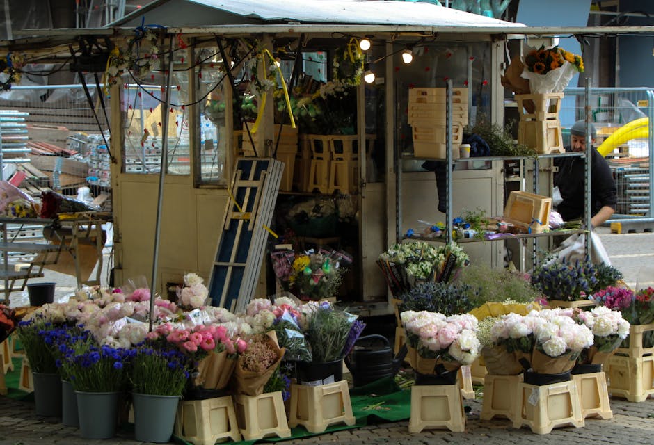 A busy outdoor flower stall displaying a vibrant assortment of fresh blooms arranged in black buckets and wrapped in brown paper, including pink peonies, white roses, purple lavender, and blue irises.